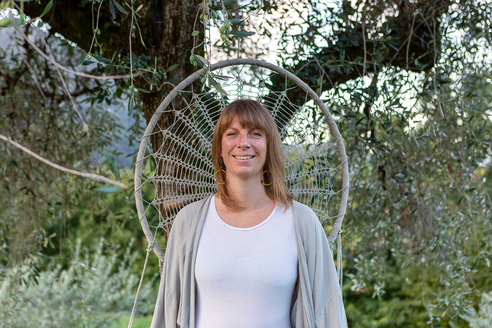 Nastassja Ugolini smiling with a beige fabric mandala and trees background, offering de-armoring and somatic bodywork